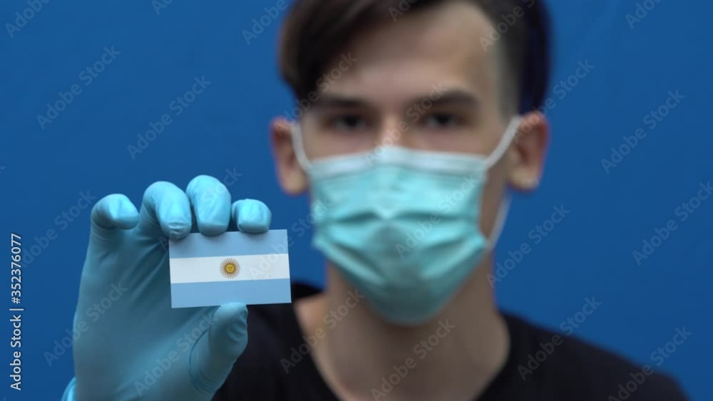 COVID-19 pandemic in Argentina. A young, attractive man, Latin American, Argentinean, wearing a protective medical mask and gloves. The guy is holding the flag of Argentina. South America COVID-19 pandemic in Argentina. A young, attractive man, Latin American, Argentinean, wearing a protective medical mask and gloves. The guy is holding the flag of Argentina. South America