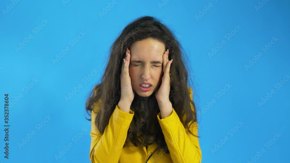 Beautiful girl is suffering from a migraine standing on a blue background, slow motion Beautiful girl is suffering from a migraine standing on a blue background, slow motion