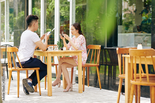 Young Asian couple checking social media and texting friends when having breaksfast in outdoor cafe
