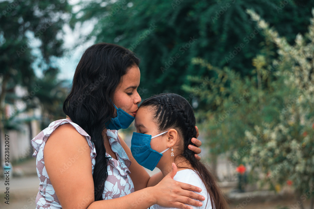 Mamá con mascarilla besando a su hija en la frente en el día de la madre al aire libre Mamá con mascarilla besando a su hija en la frente en el día de la madre al aire libre