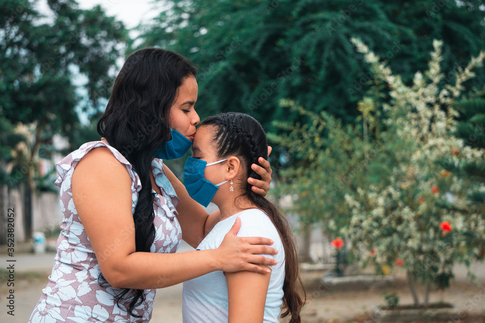 Mamá con mascarilla besando a su hija en la frente en el día de la madre al aire libre Mamá con mascarilla besando a su hija en la frente en el día de la madre al aire libre