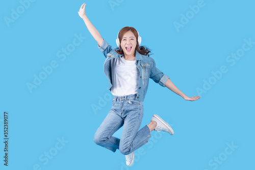 Beautiful Asian woman is smiling happily while listening to music and jumping. On a blue background in the studio.