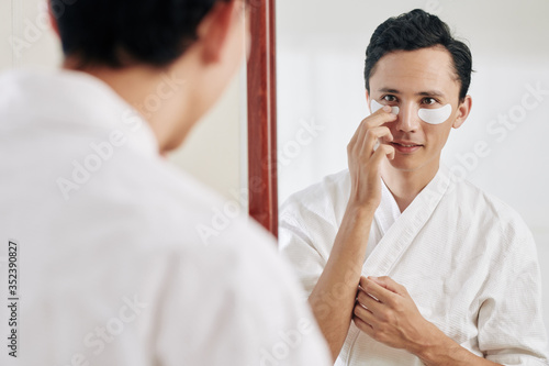 Smiling man in waffle bath robe looking at mirror and applying eye patches