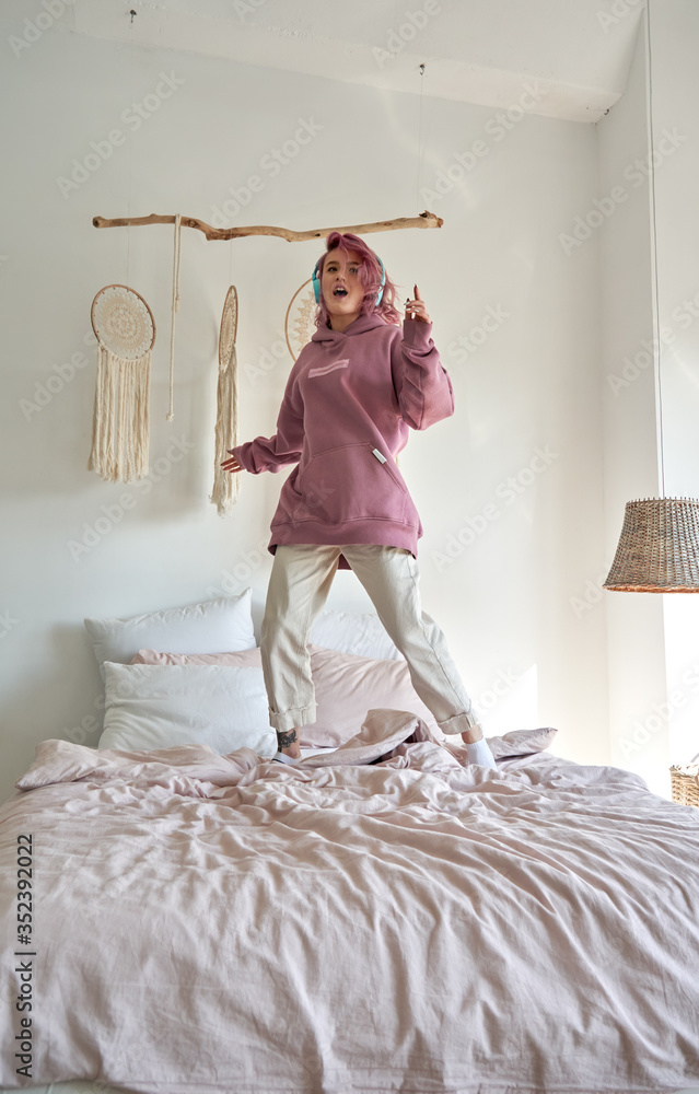 Happy funny hipster teenage girl with pink hair wear hoodie and headphones listening radio pop music playlist podcast, singing song jumping dancing standing on bed in cozy hygge bedroom. Vertical Stock Photo | Happy funny hipster teenage girl with pink hair wear hoodie and headphones listening radio pop music playlist podcast, singing song jumping dancing standing on bed in cozy hygge bedroom. Vertical Stock Photo |