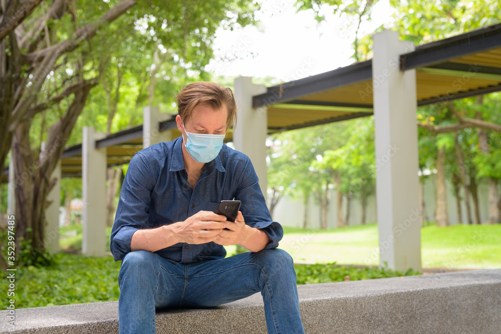 Young man with mask using phone and sitting at the park