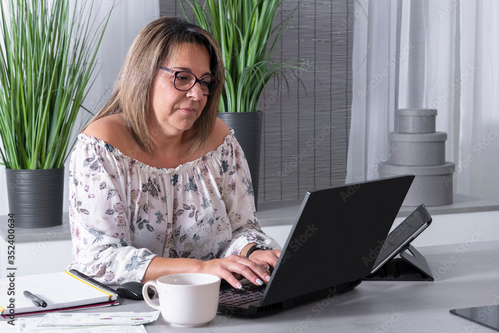 Fototapeta premium mature woman working on a laptop surrounded by a notebook, a mug and a tablet.