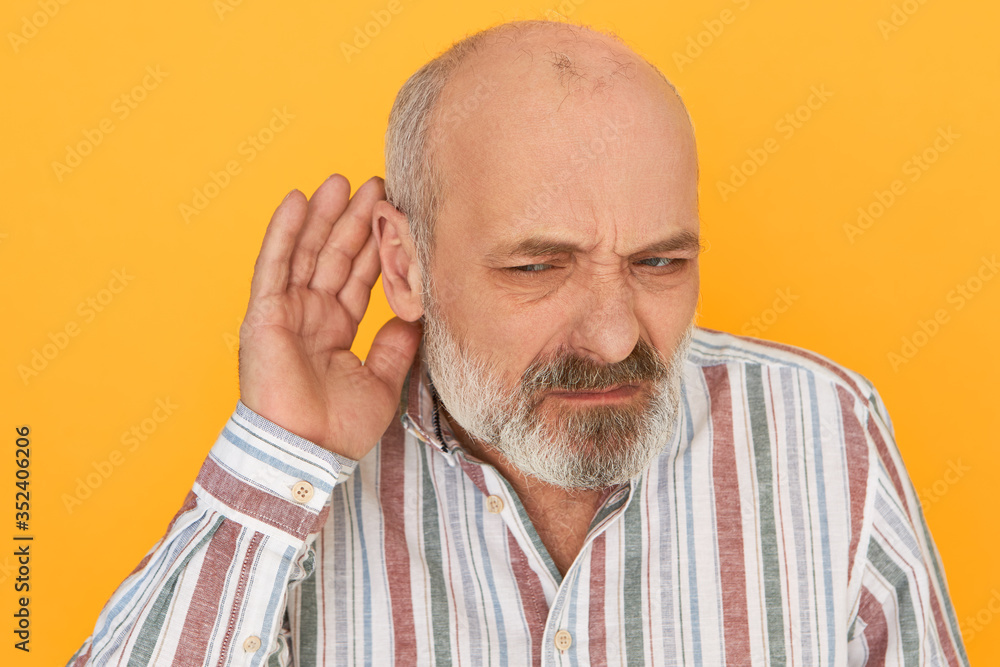 Obraz premium Portrait of frowning frustrated bearded male pensioner in striped shirt keeping hand at his ear, listening attentively, trying to hear indistinct talking. Hearing problems and eavesdropping