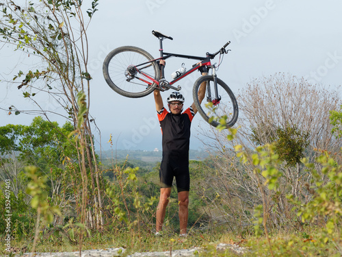 A mountain biker is happy holding his bike above his head while standing on top of a mountain with an ocean view.