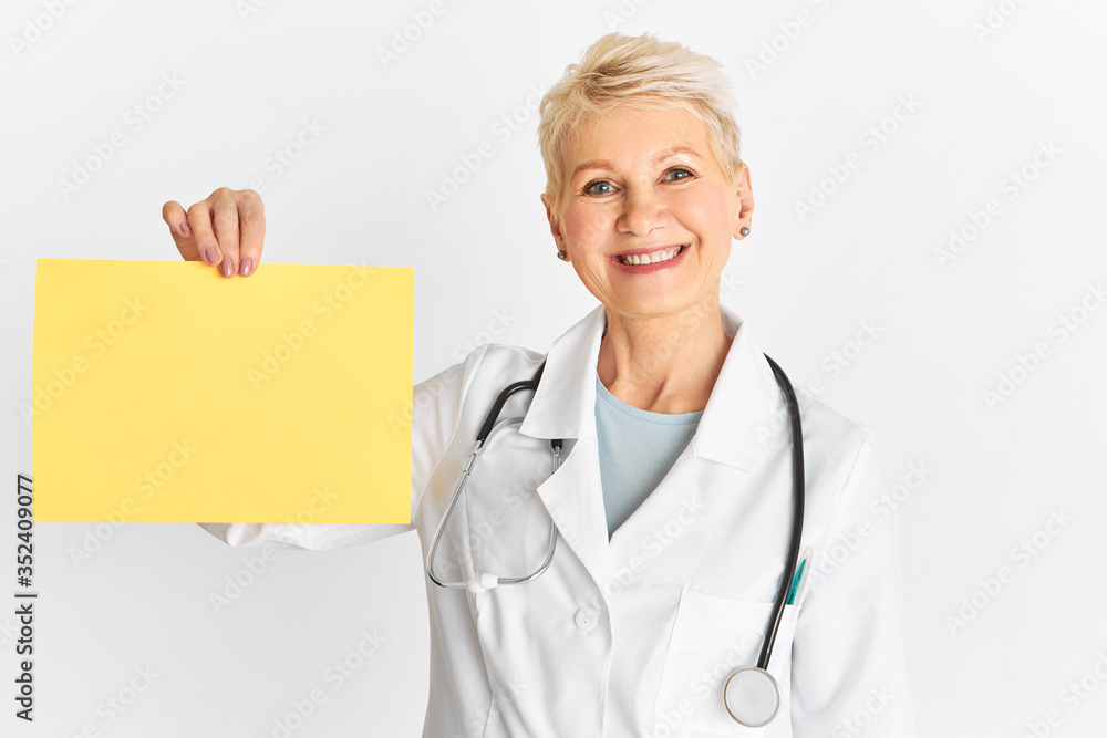 Isolated shot of good looking optimistic senior woman doctor with blonde pixie hair and cheerful confident smile holding blank yellow banner with copy space for your promotional information Isolated shot of good looking optimistic senior woman doctor with blonde pixie hair and cheerful confident smile holding blank yellow banner with copy space for your promotional information