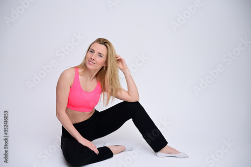 Portrait of a pretty fitness blonde girl in a sports uniform on a white background sitting on the floor. A slender Caucasian model poses with a smile.