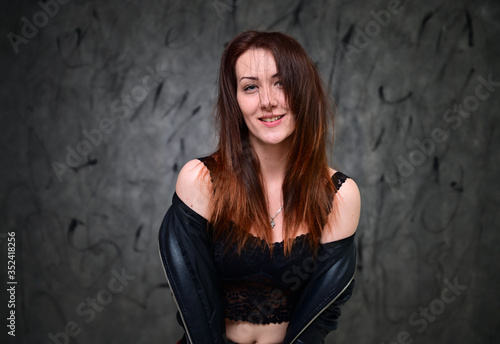 Studio portrait of a caucasian young brunette woman with minimal makeup. A pretty model is standing on a gray background in a black jacket.