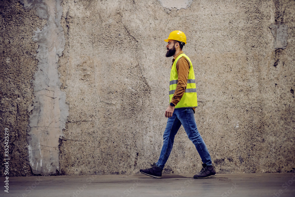 Side view of handsome hardworking bearded worker in vest, with safety helmet on head passing by concrete wall in building in construction process.