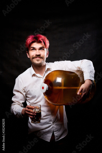 Young handsome guy with red hair in white shirt with beer. Funny man with large bottle of beer and black background. Brewer and barrel