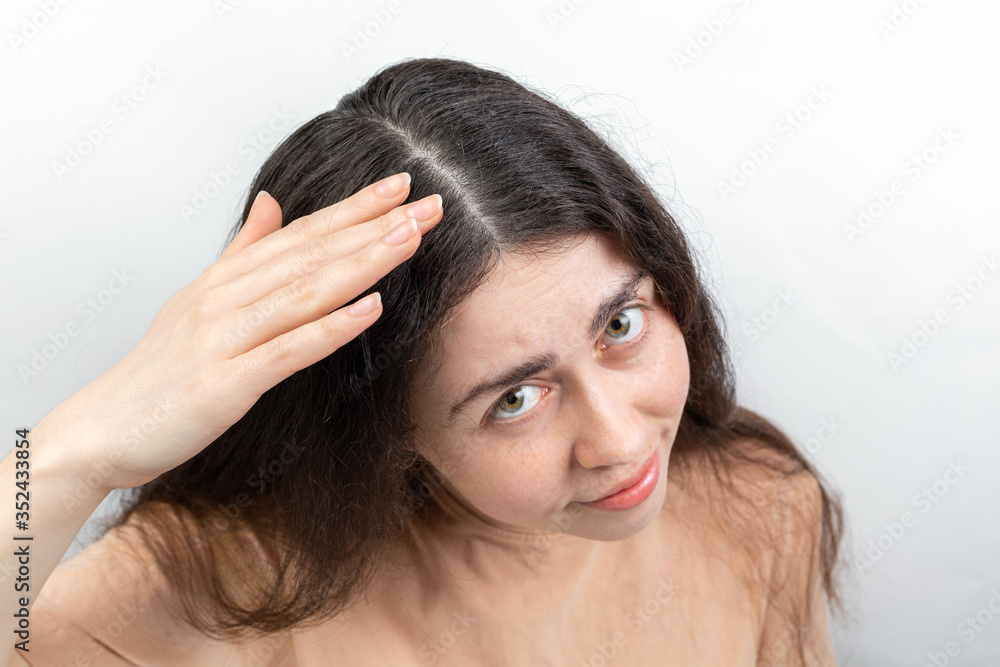 Naklejka premium Dandruff and seborrhea. Portrait of a young woman with dark hair, anxiously brushing dandruff from her hair. Gray background. Concept of problems with hair and head lice