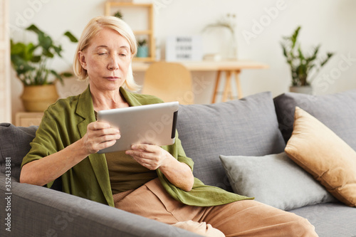 Mature woman in casual clothing sitting on sofa and watching something on digital tablet in the living room at home