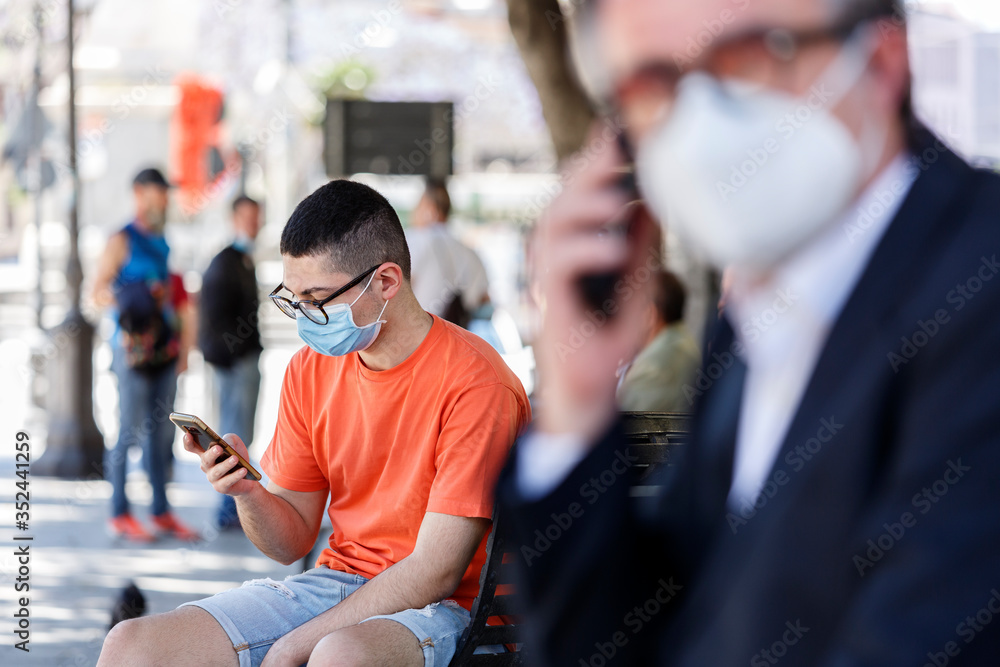 ragazzo con occhiali neri e maglia arancione indossa un mascherina chirurgica e guarda il suo cellulare seduto in una panchina nella piazza del centro di una città ragazzo con occhiali neri e maglia arancione indossa un mascherina chirurgica e guarda il suo cellulare seduto in una panchina nella piazza del centro di una città