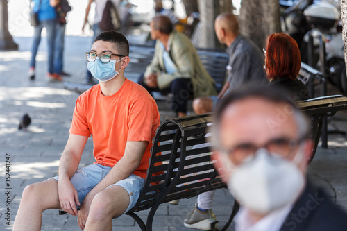 ragazzo con occhiali neri e maglia arancione indossa un mascherina chirurgica seduto  in una panchina nella piazza del centro di una città  photo