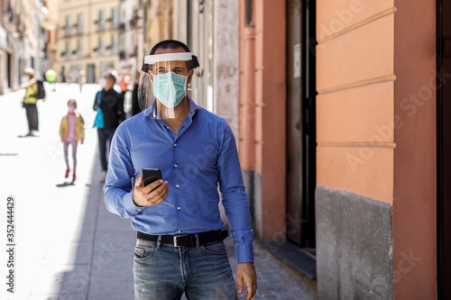 Uomo con camicia blu e mascherina facciale protettiva in mezzo al marciapiede di una città con il cellulare in mano e con persone in movimento introno © alex.pin