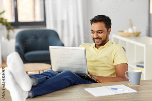 technology, remote job and lifestyle concept - happy smiling indian man with laptop computer resting feet on table at home office