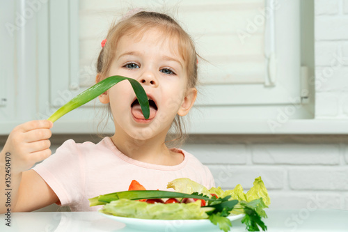 4 years old girl sits in the kitchen at the table and eats vegetables, bites green onions. preschool nutrition rules