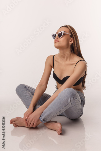 portrait of sexy asian woman with long hair posing in black lingerie, sunglasses, jeans on white studio background with bare feet. model tests of skinny girl in bra. attractive female sitting on floor