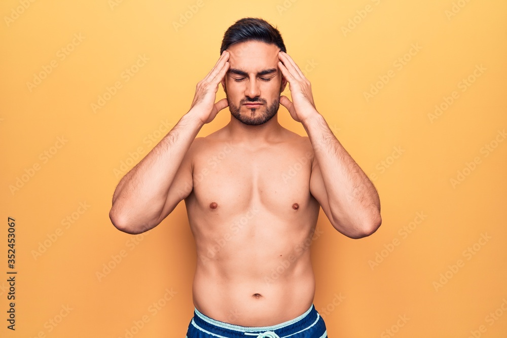 Young handsome man with beard wearing sleeveless t-shirt standing over yellow background suffering from headache desperate and stressed because pain and migraine. Hands on head.