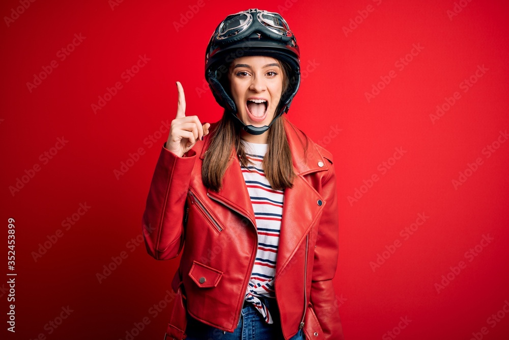 Young beautiful brunette motocyclist woman wearing motorcycle helmet and red jacket pointing finger up with successful idea. Exited and happy. Number one. Young beautiful brunette motocyclist woman wearing motorcycle helmet and red jacket pointing finger up with successful idea. Exited and happy. Number one.