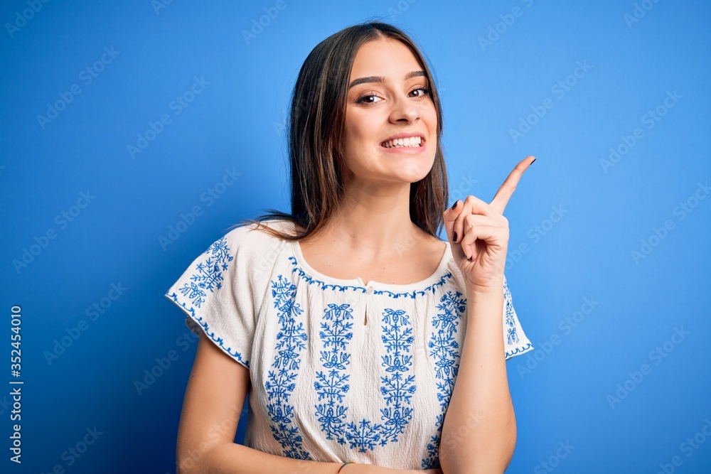 Young beautiful brunette woman wearing casual t-shirt standing over blue background with a big smile on face, pointing with hand and finger to the side looking at the camera. Young beautiful brunette woman wearing casual t-shirt standing over blue background with a big smile on face, pointing with hand and finger to the side looking at the camera.