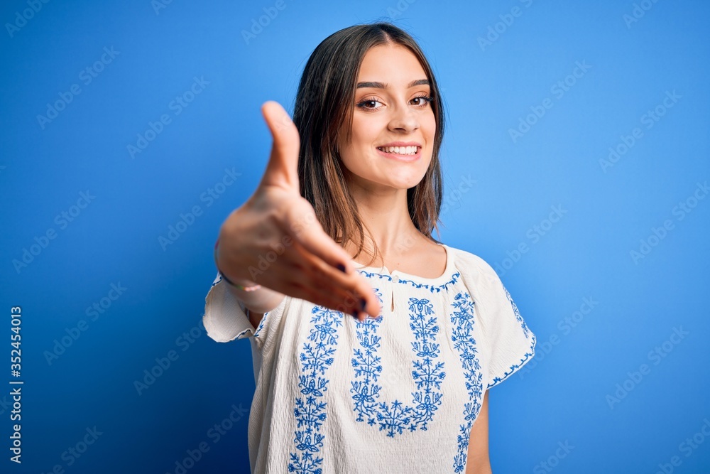 Young beautiful brunette woman wearing casual t-shirt standing over blue background smiling friendly offering handshake as greeting and welcoming. Successful business. Young beautiful brunette woman wearing casual t-shirt standing over blue background smiling friendly offering handshake as greeting and welcoming. Successful business.