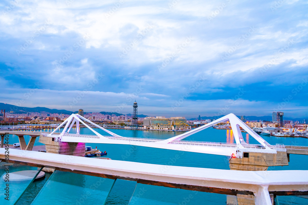 Barcelona, Spain. Porta d’Europa bascule mobile bridge with Torre Jaume I aerial lift pylon steel truss tower, mountains and city skyline architecture in background. Barcelona, Spain. Porta d’Europa bascule mobile bridge with Torre Jaume I aerial lift pylon steel truss tower, mountains and city skyline architecture in background.