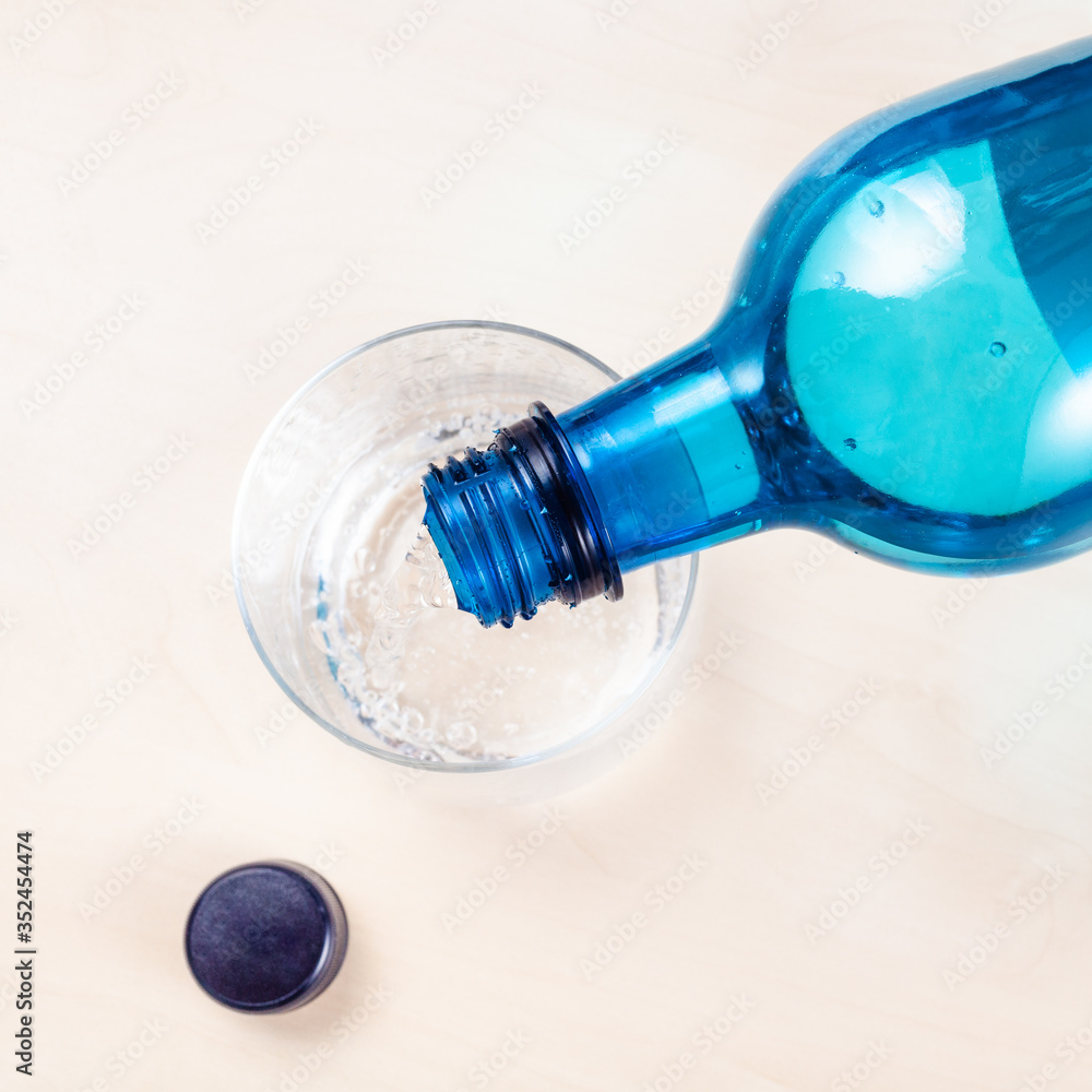 top view of pouring sparkling mineral water from blue plastic bottle in glass on light brown table close up top view of pouring sparkling mineral water from blue plastic bottle in glass on light brown table close up