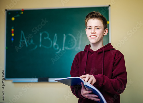 Student of higer school is standing in dark hoodie near the green blackboard in the classroom reading notes. Front view. School education concept.