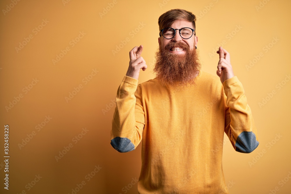 Naklejka premium Handsome Irish redhead man with beard wearing glasses over yellow isolated background gesturing finger crossed smiling with hope and eyes closed. Luck and superstitious concept.