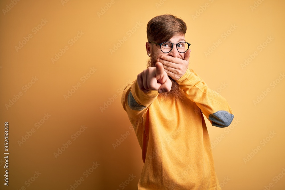Handsome Irish redhead man with beard wearing glasses over yellow isolated background laughing at you, pointing finger to the camera with hand over mouth, shame expression Handsome Irish redhead man with beard wearing glasses over yellow isolated background laughing at you, pointing finger to the camera with hand over mouth, shame expression