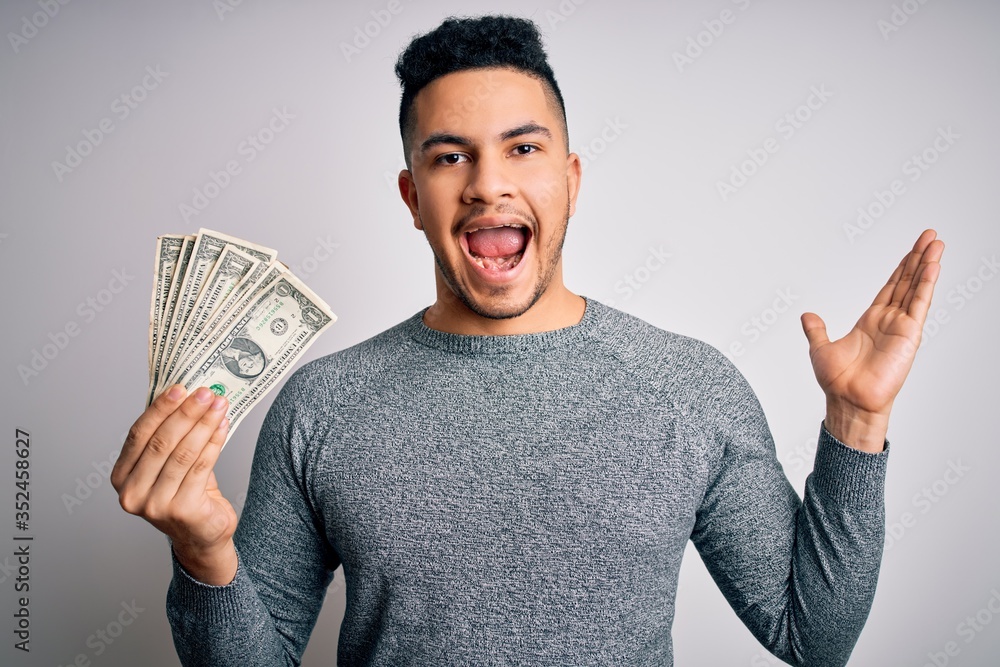 Young handsome man holding bunch of dollars banknotes over isolated white background very happy and excited, winner expression celebrating victory screaming with big smile and raised hands Young handsome man holding bunch of dollars banknotes over isolated white background very happy and excited, winner expression celebrating victory screaming with big smile and raised hands