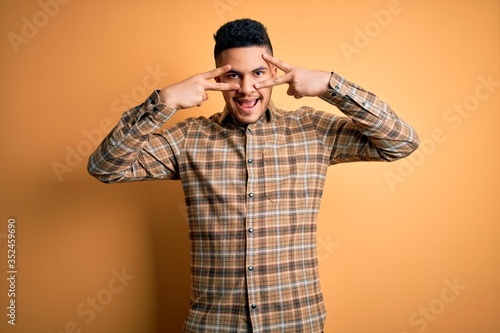 Young handsome man wearing casual shirt standing over isolated yellow background Doing peace symbol with fingers over face, smiling cheerful showing victory
