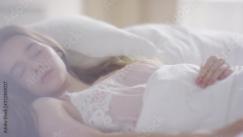  Slowmo PAN of beautiful young woman in white nightdress sleeping in bed on peaceful morning, then waking up and smiling