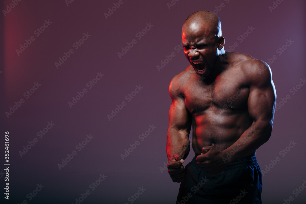 A professional dark-skinned boxer demonstrates the muscles of his torso and makes a war cry against a neon red background.