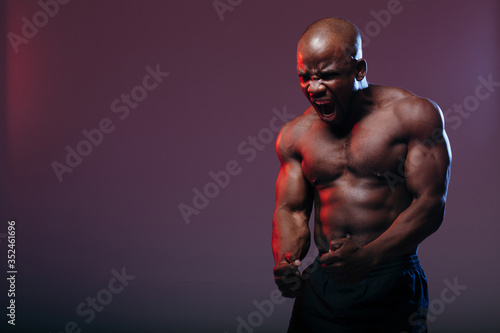 A professional dark-skinned boxer demonstrates the muscles of his torso and makes a war cry against a neon red background.