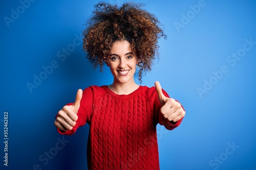 Young beautiful woman with curly hair and piercing wearing casual red sweater approving doing positive gesture with hand, thumbs up smiling and happy for success. Winner gesture.