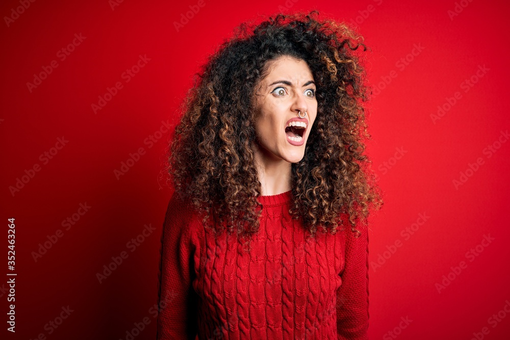 Young beautiful woman with curly hair and piercing wearing casual red sweater angry and mad screaming frustrated and furious, shouting with anger. Rage and aggressive concept. Young beautiful woman with curly hair and piercing wearing casual red sweater angry and mad screaming frustrated and furious, shouting with anger. Rage and aggressive concept.