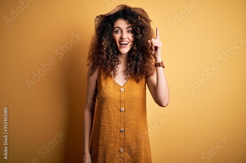 Beautiful brunette woman on vacation with curly hair and piercing wearing hat and dress pointing finger up with successful idea. Exited and happy. Number one.