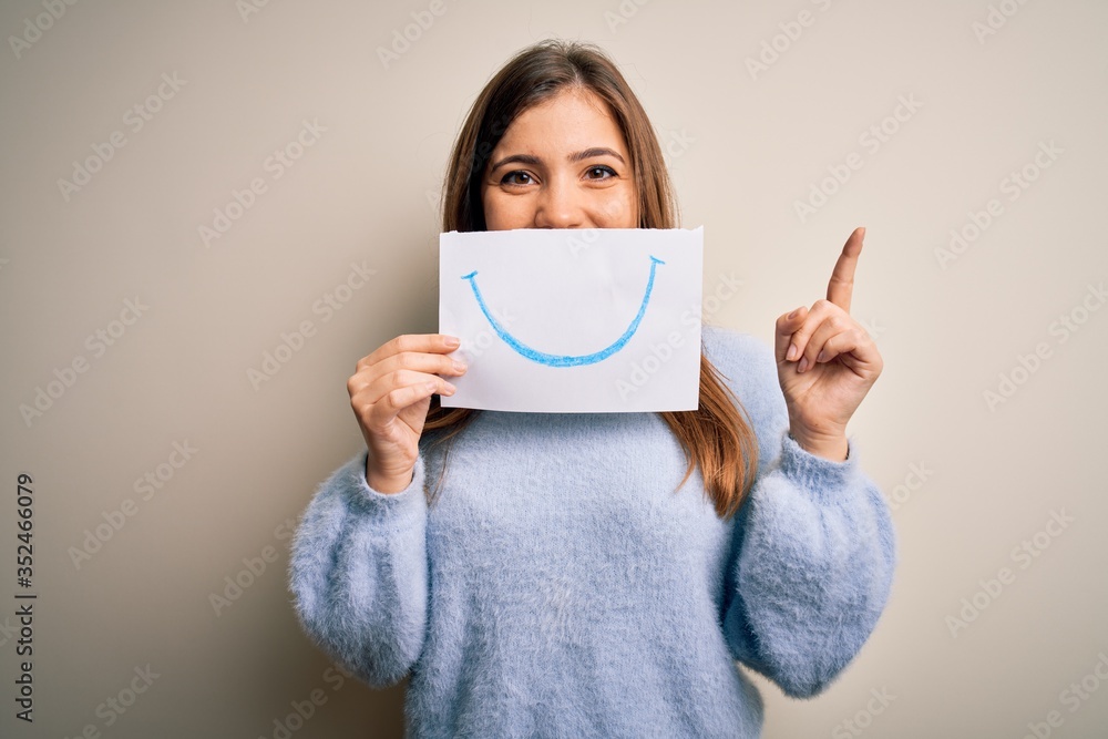 Young blonde woman holding funny smile drawing on mouth as happy expression surprised with an idea or question pointing finger with happy face, number one Young blonde woman holding funny smile drawing on mouth as happy expression surprised with an idea or question pointing finger with happy face, number one