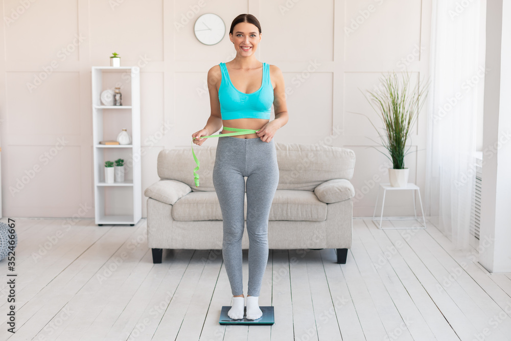 Smiling Girl Measuring Thin Waist Standing On Weight-Scales At Home Smiling Girl Measuring Thin Waist Standing On Weight-Scales At Home
