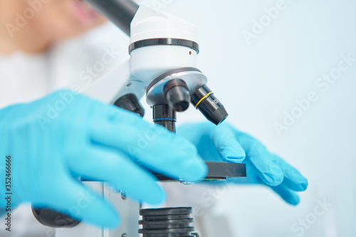 Close-up of female lab technician in blue rubber gloves looking through microscope