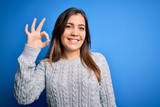Beautiful young woman wearing casual wool sweater standing over blue isolated background smiling positive doing ok sign with hand and fingers. Successful expression.