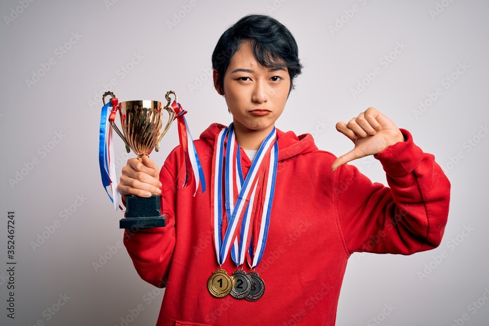Young beautiful asian girl winner holding trophy wearing medals over white background with angry face, negative sign showing dislike with thumbs down, rejection concept Young beautiful asian girl winner holding trophy wearing medals over white background with angry face, negative sign showing dislike with thumbs down, rejection concept