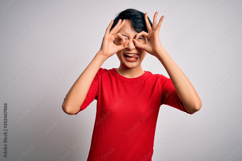 Young beautiful asian girl wearing casual red t-shirt standing over isolated white background doing ok gesture like binoculars sticking tongue out, eyes looking through fingers. Crazy expression. Young beautiful asian girl wearing casual red t-shirt standing over isolated white background doing ok gesture like binoculars sticking tongue out, eyes looking through fingers. Crazy expression.