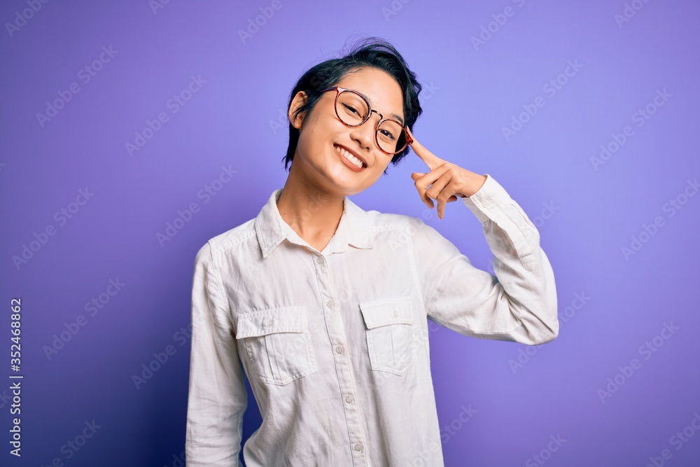Young beautiful asian girl wearing casual shirt and glasses standing over purple background Smiling pointing to head with one finger, great idea or thought, good memory Young beautiful asian girl wearing casual shirt and glasses standing over purple background Smiling pointing to head with one finger, great idea or thought, good memory