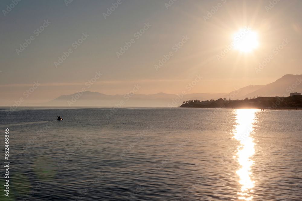 Silhouette of a man on a kayak against the backdrop of the setting sun with a bright glare on the sea on the background of the coast with palm trees and mountains